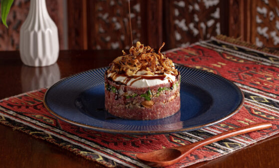 Prato Kibe Burj montado em formato de torre, com base de kibe cru bem temperado, camada de carne moída (parte crua e parte cozida com amêndoas e melaço de romã), tabule fresco com salsa e tomate, cobertura de coalhada seca e finalização com cebola crocante. A apresentação destaca o contraste entre o vermelho do kibe, o verde do tabule, o branco da coalhada e os tons dourados da cebola e das amêndoas.