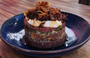 Prato Kibe Burj montado em formato de torre, com base de kibe cru bem temperado, camada de carne moída (parte crua e parte cozida com amêndoas e melaço de romã), tabule fresco com salsa e tomate, cobertura de coalhada seca e finalização com cebola crocante. A apresentação destaca o contraste entre o vermelho do kibe, o verde do tabule, o branco da coalhada e os tons dourados da cebola e das amêndoas.